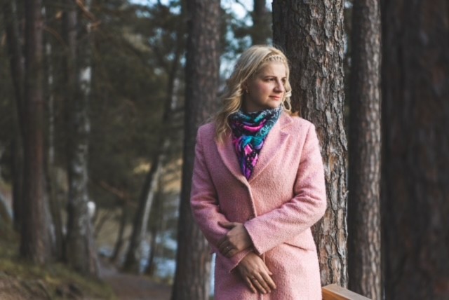 Nainen vaaleanpunaisessa takissa seisoo syksyisessä metsässä ja katsoo sivulle.