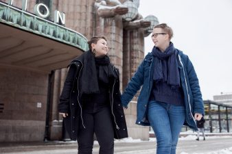 Kaksi helsinkiläistä hymyilee ja kävelee kädet kädessä Helsingin rautatieaseman edessä.